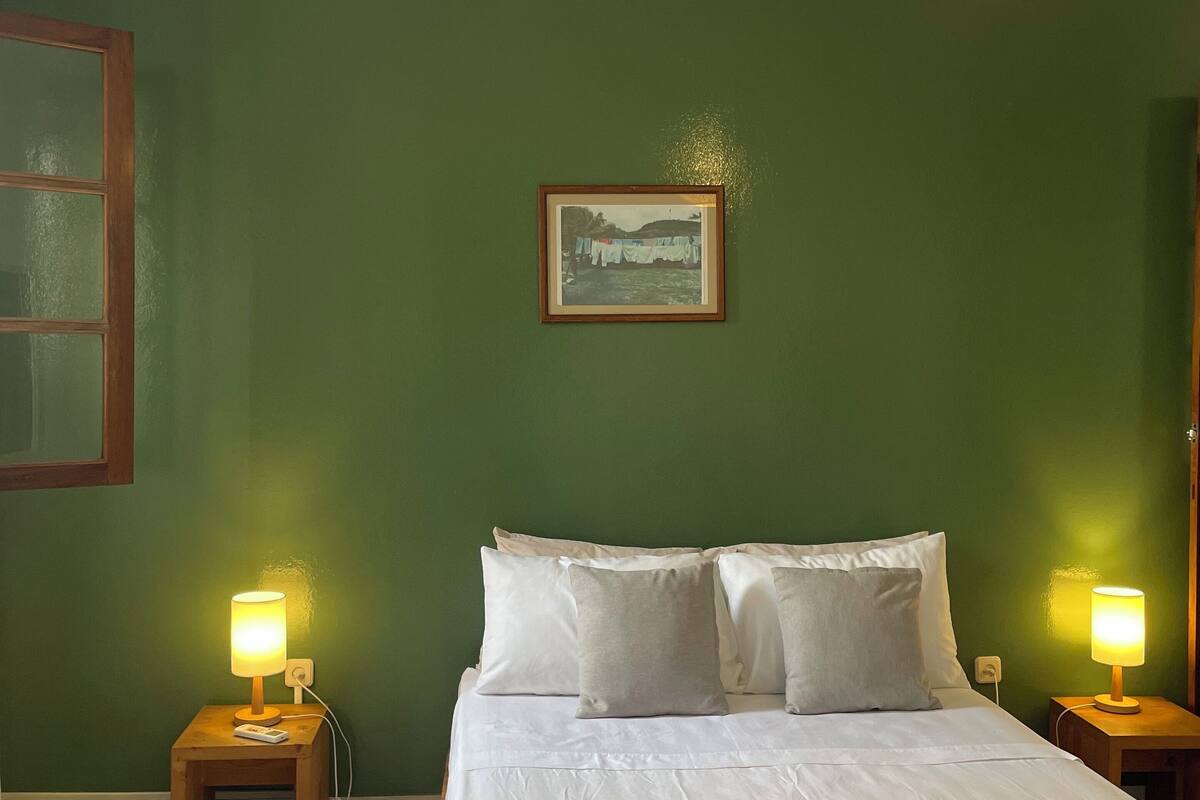 A bedroom with a bed, two bedside lamps, a hanging light fixture, a framed picture on the wall, and a window with a wooden frame.