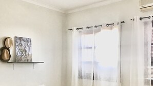 A bedroom with a bed, white curtains, a painting on the wall, and a shelf with two hats. - Syds Guesthouse I (Praia)
