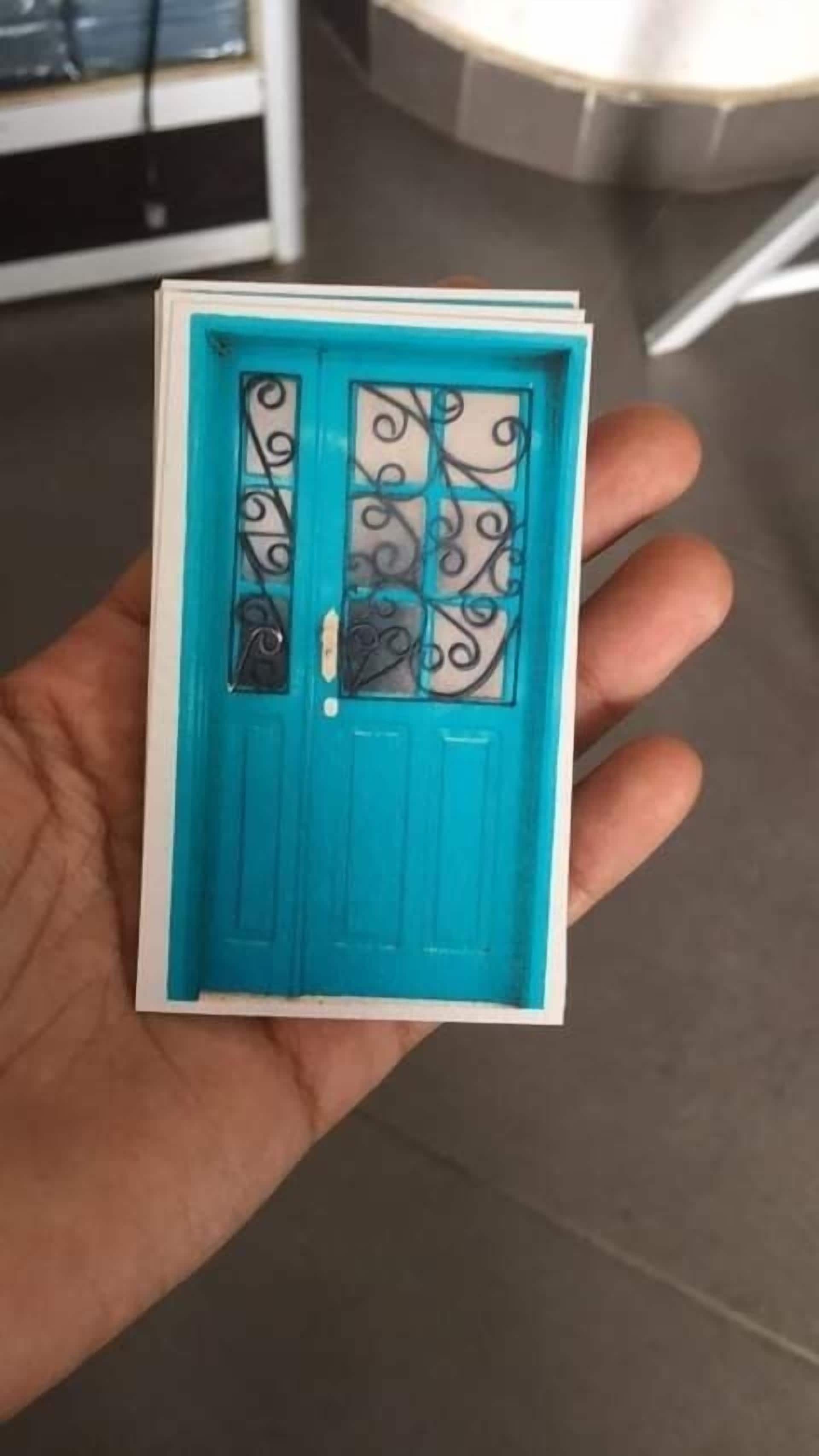 Una mano sostiene una maqueta de una puerta azul con ornamentación de hierro.