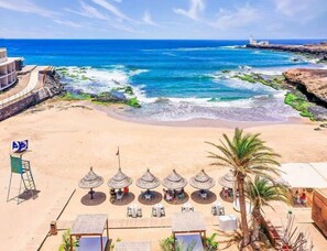 Una playa con sombrillas y mesas, una torre de salvavidas y un faro al fondo.