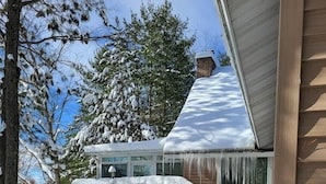 Una casa de madera con tejado nevado y una terraza de madera que da a una puerta de cristal.