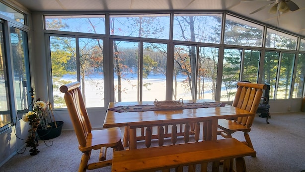 Una habitación iluminada por el sol con un ventanal que da a un paisaje nevado, una mesa de madera con sillas y una lámpara.