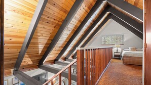 Ein Schlafzimmer im Loft-Stil mit schrägem Dach, Holzbalken, Teppichboden, einem Bett, einem Nachttisch und einem Fenster mit Jalousien.
