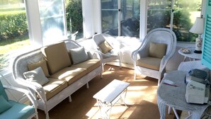 Une pièce baignée de lumière naturelle, meublée de meubles en osier, dotée de grandes fenêtres et comprenant une petite table sur laquelle se trouve une imprimante.