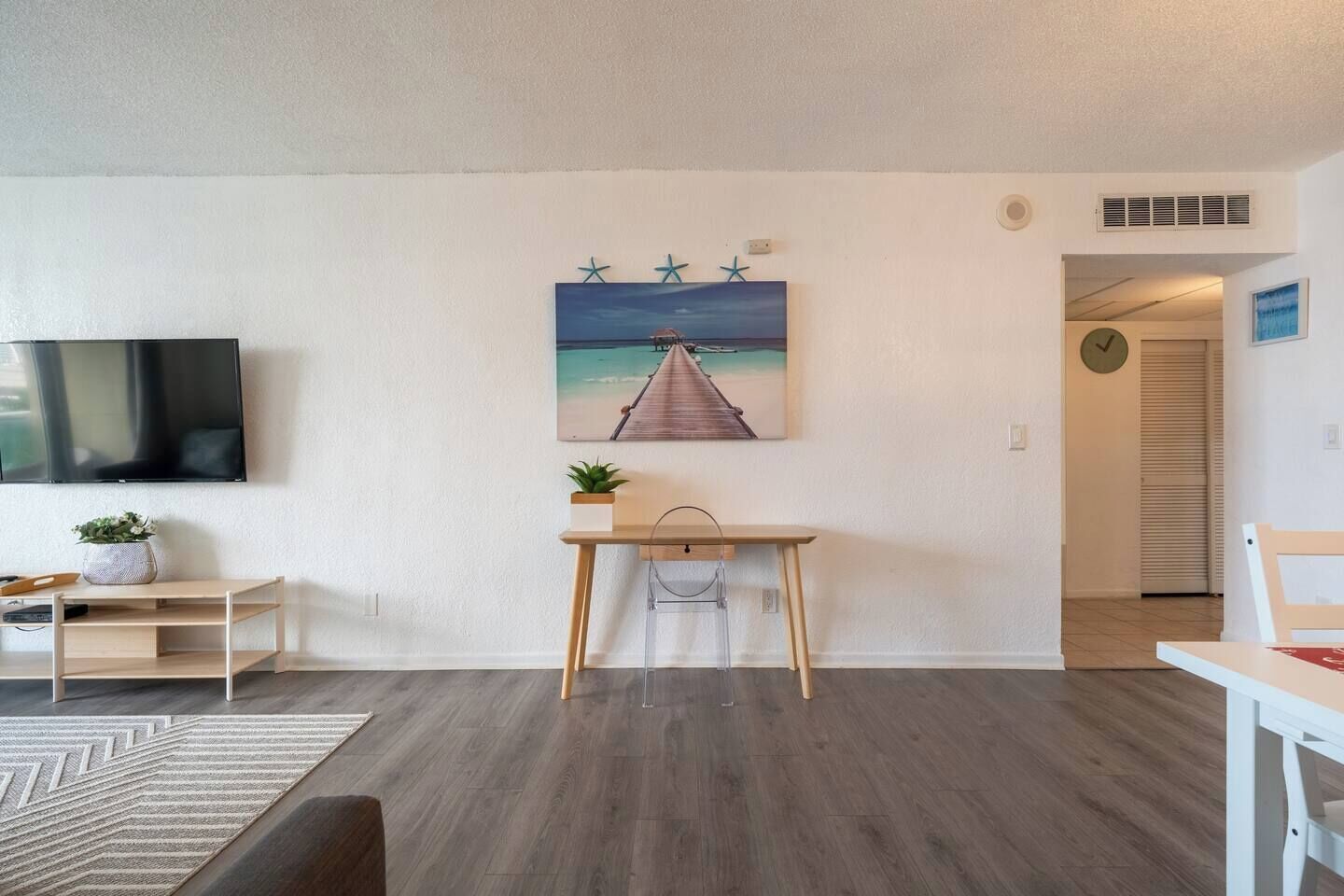 Un moderno salón con piso de madera, un televisor de pantalla plana en la pared, un escritorio con una planta y un cuadro de un muelle.