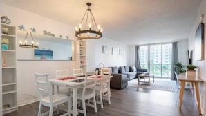 A modern living room with a dining area, a grey sofa, and a large window with a view of buildings. - Beautiful 1 bedroom apartment fully furnished steps away from the beach (Sunny Isles Beach)