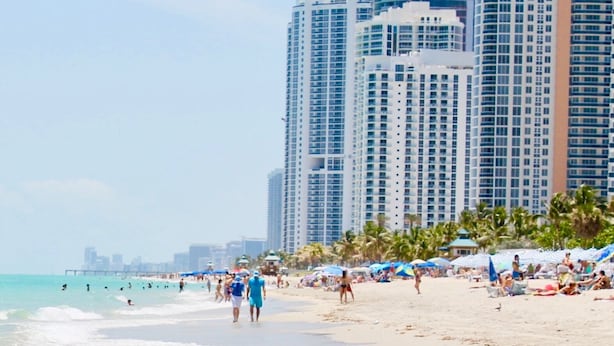 A beach with people walking along the shore, palm trees, and tall buildings in the background. - Beautiful 1 bedroom apartment fully furnished steps away from the beach (Sunny Isles Beach)
