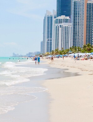 A beach with people walking along the shore, palm trees, and tall buildings in the background.