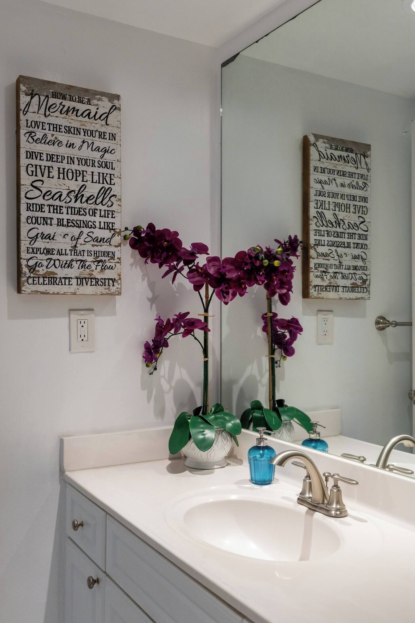 A bathroom with a white sink, two purple orchids, and two framed inspirational quotes on the wall.