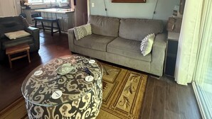 Un soggiorno con un divano grigio, un poggiapiedi decorato, una zona pranzo con tavolo e sedie, una cucina sullo sfondo e un quadro appeso al muro.