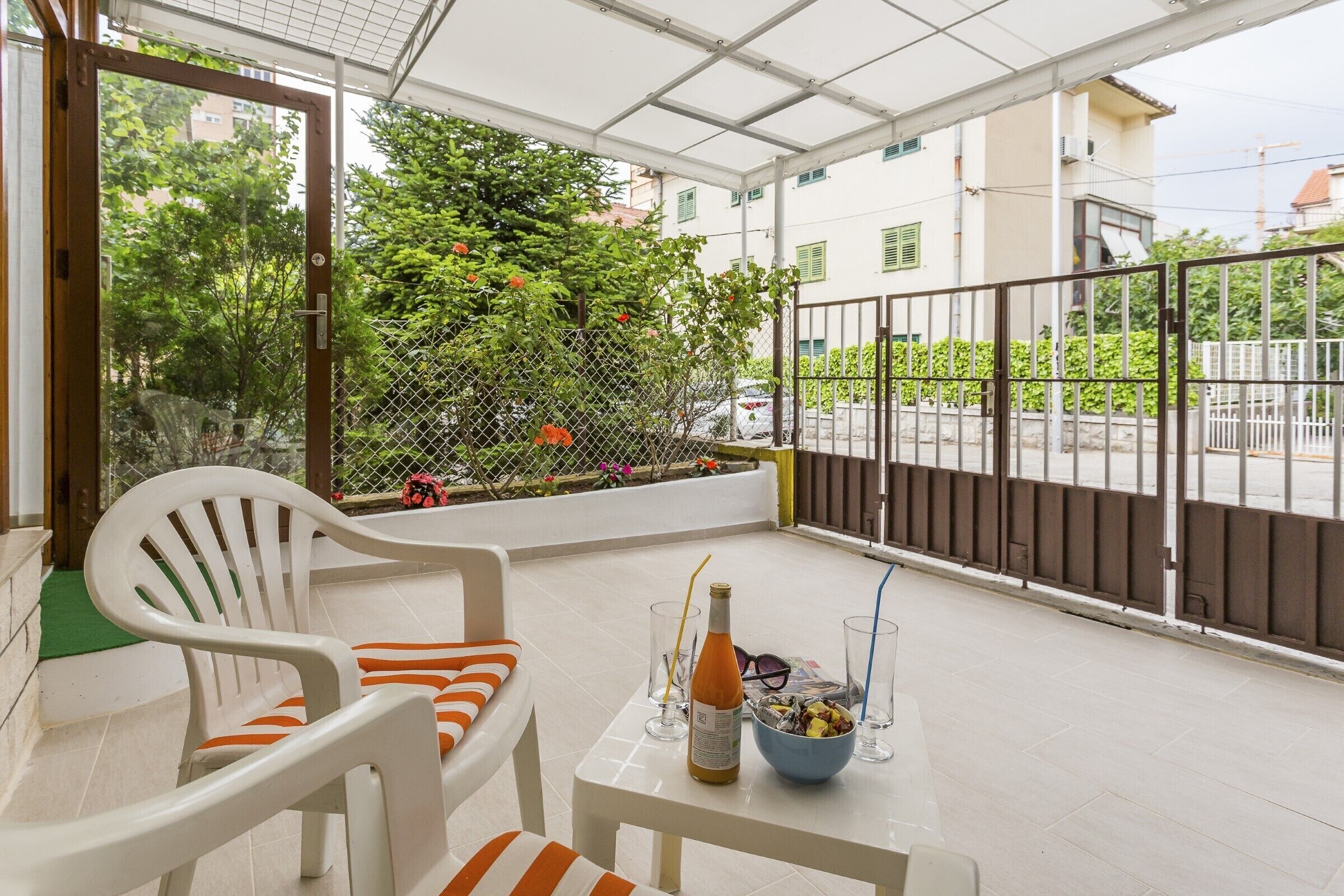 Ein Balkon mit weißem Stuhl, einem Tisch mit Flasche und Gläsern, und Blick auf eine Straße mit Gebäuden und Grünflächen.