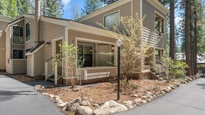 A two-story building with a covered porch, a staircase, and a small garden area with rocks and young trees. - Raccoon Roost Condo | Community Pool & Club House (Incline Village)