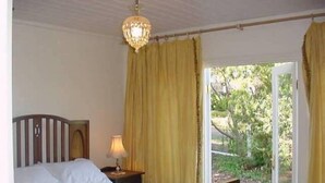 Un dormitorio con cama, mesita de noche, lámpara y vistas al verde exterior.