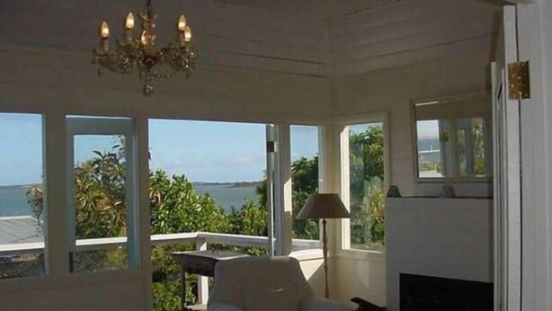 Una habitación con un sillón blanco, una mesa, una lámpara y una chimenea. Amplias ventanas ofrecen vistas al mar y la vegetación.