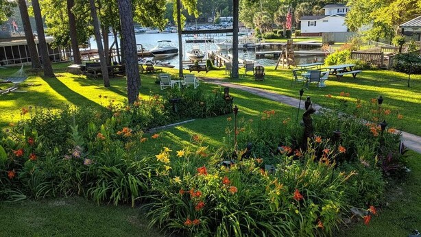 Ein gepflegter Garten mit verschiedenen Blumen und Bäumen, ein Picknickplatz und eine Aussicht auf Boote auf dem Wasser.