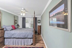 A bedroom with a four-poster bed, a ceiling fan, a large framed picture, and a view of the outdoors through a window.