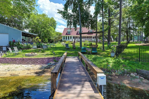 A wooden bridge leading to a house with a porch, surrounded by a grassy yard with chairs and a small pond.