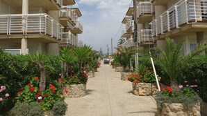 A pathway flanked by multi-story buildings with balconies, stone planters with red flowers, and a clear sky. - Large apartment. Front line beach, private parking, bicycles (Peñiscola)