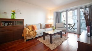 A modern living room with a sofa, a coffee table, and a chair. There is a wooden cabinet with books and a vase with flowers. A large window with curtains is visible. - Two bedroom apartment with terrace - You Stylish (Barcelona)