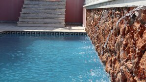 Una piscina con agua azul cristalina, un muro de piedra y una escalera que lleva a un edificio con barandilla blanca.