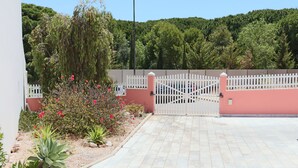 Una entrada con reja y cerca de piquetes blancos, camino de entrada pavimentado y diversos plantas y arbustos.