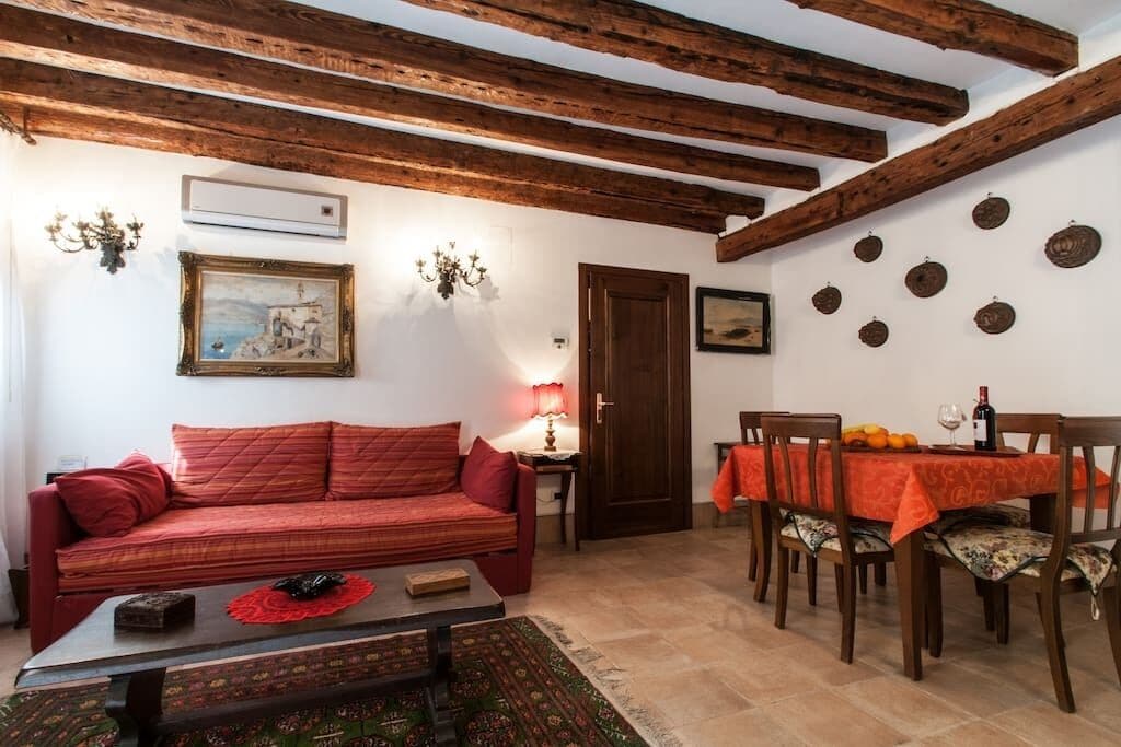 A cozy living room with a red sofa, wooden beams on the ceiling, a dining table with chairs, and a small table with a lamp.