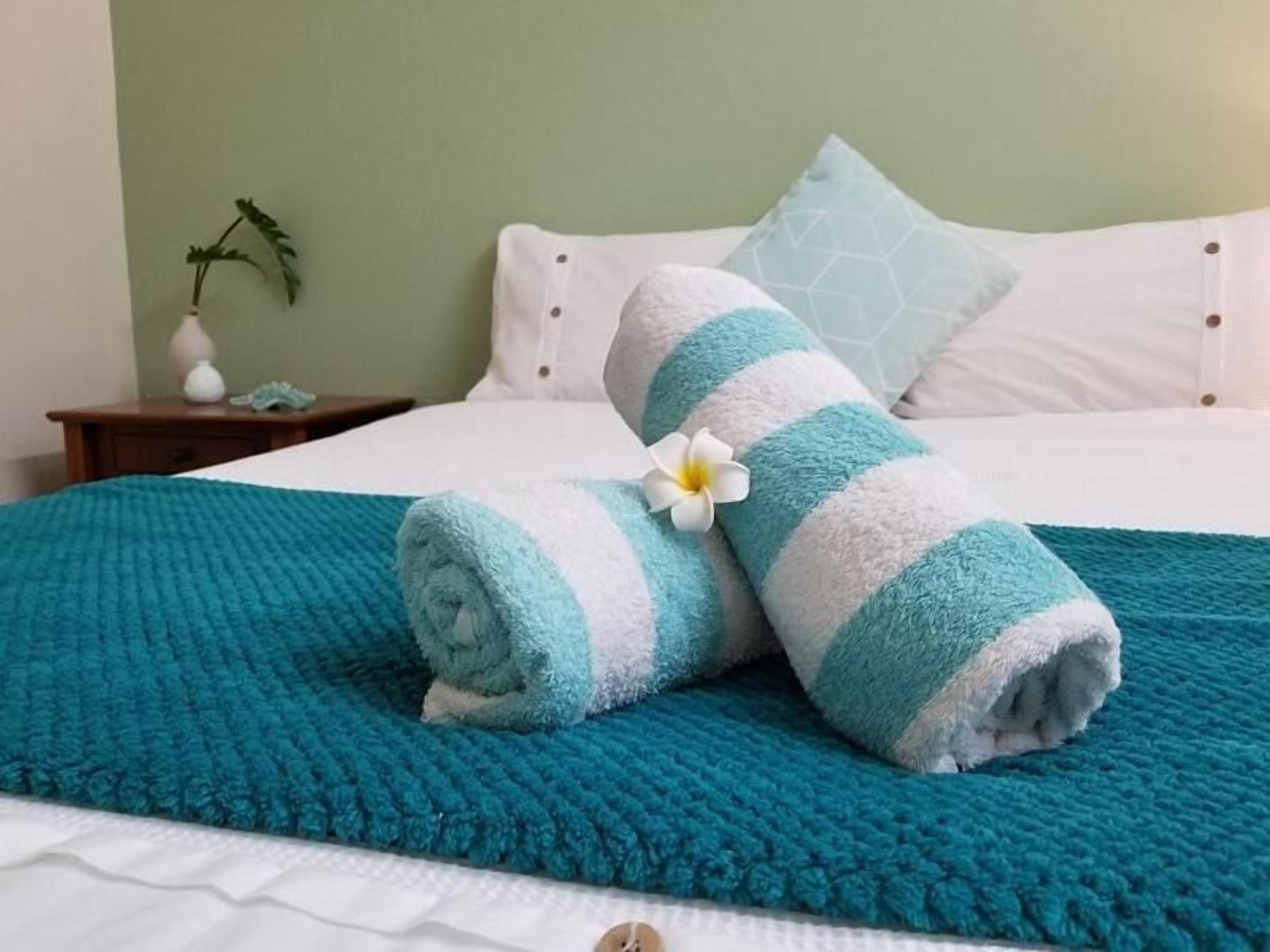 A neatly made bed with a teal blanket, a rolled towel with a white flower, and a small nightstand with a vase and a plant.