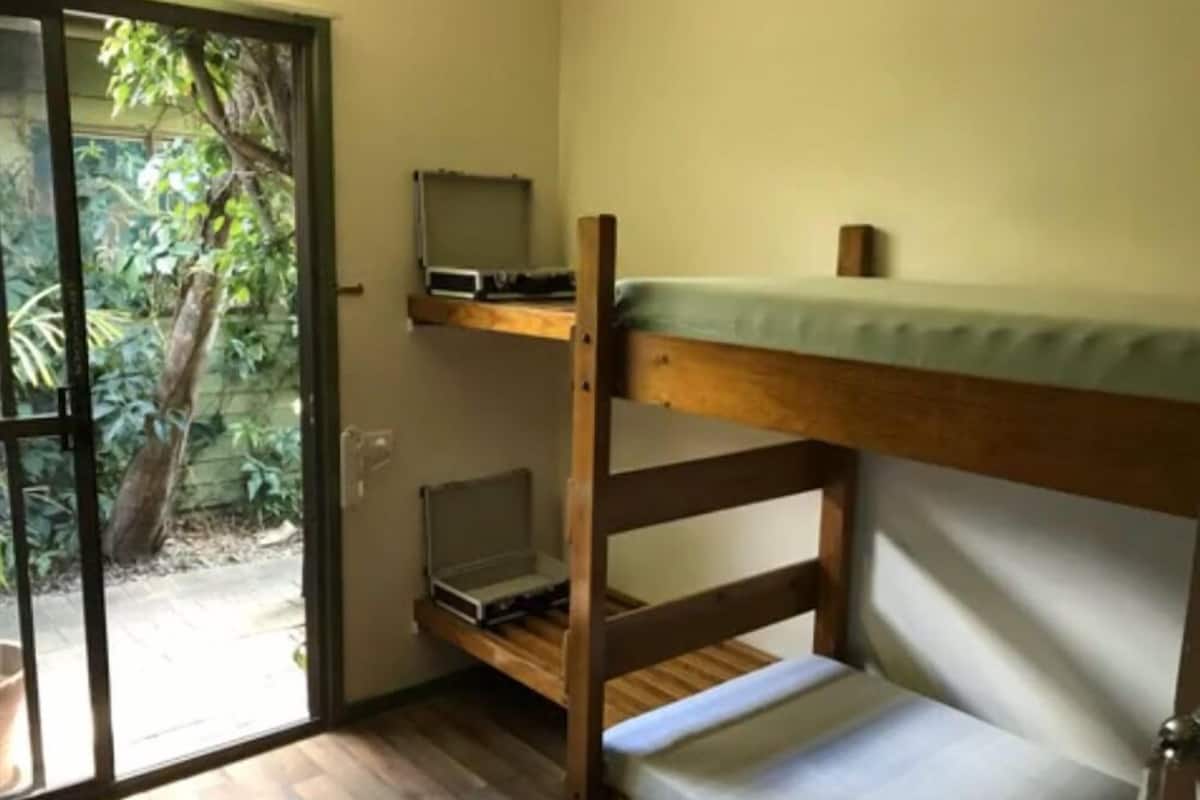Una litera con una computadora portátil en el estante inferior, una mesita pequeña y una puerta corrediza de vidrio que da a un jardín.