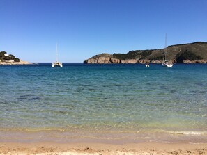 Una playa con agua cristalina, una orilla arenosa y dos veleros anclados cerca de acantilados rocosos.