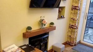 Een woonkamer met een open haard, een aan de muur bevestigde televisie, een klein houten tafeltje en een bank.
