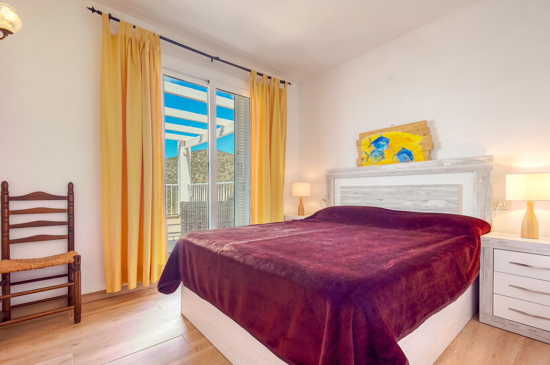 A bedroom with a bed, a chair, a nightstand, a lamp, a painting, and a view of the outdoors.