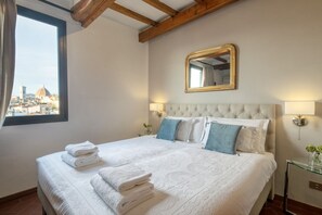 A hotel room with a bed, a window with a city view, a mirror, and wall-mounted lamps.