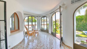 Un couloir baigné de lumière, avec des carreaux de sol à motifs, un fauteuil en osier et de grandes fenêtres en arc qui offrent une vue sur un jardin et une piscine.