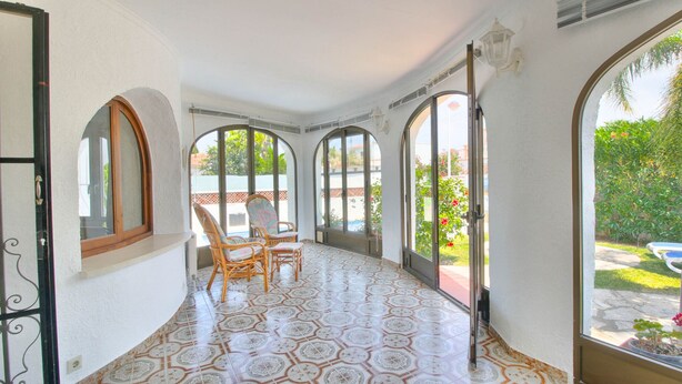 Ein sonnendurchfluteter Gang mit gemusterten Bodenfliesen, einem Korbstuhl und großen Bogenfenstern, die einen Blick auf einen Garten und ein Schwimmbecken bieten.