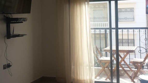 A room with a flat-screen TV mounted on the wall, a sliding glass door leading to an outdoor area with a wooden table and chairs, and a balcony with a metal railing. - Nice three rooms nine modern and well equipped (agadir)