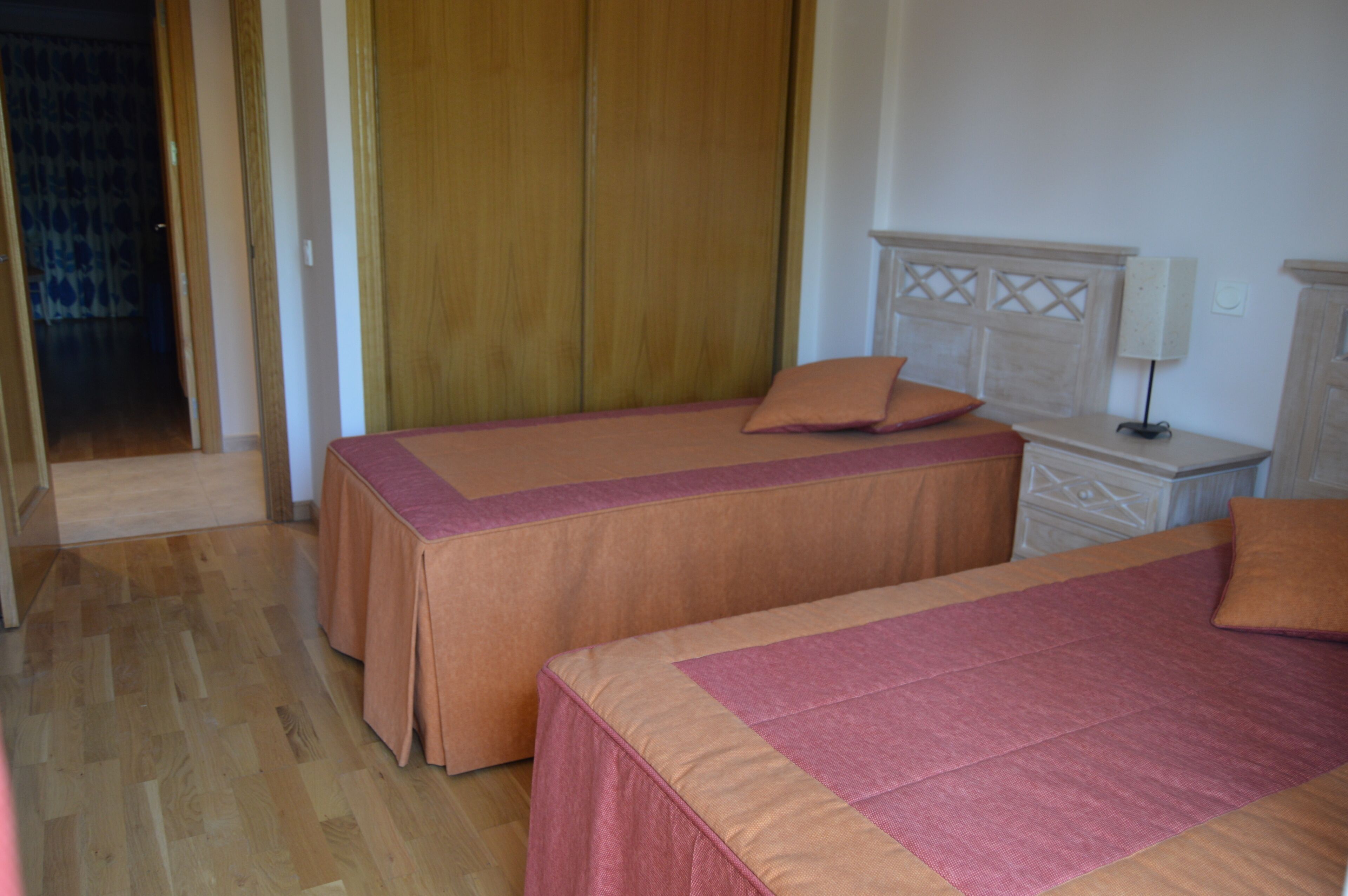 Una habitación con dos camas, un armario de madera, una mesita de noche con lámpara y una puerta que da a otra estancia.