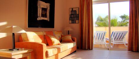 A living room with a sofa, a table, a lamp, and a balcony with chairs.