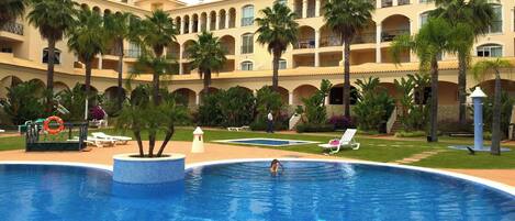 A pool area with a large pool, palm trees, and a building with balconies in the background.