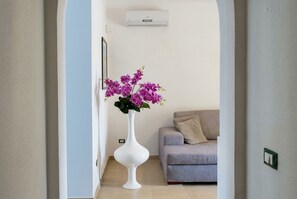A hallway with a white vase of purple flowers, a grey sofa, and an air conditioning unit on the wall.