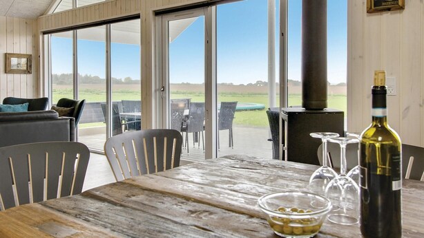 Una mesa de madera con una botella de vino y copas, un sofá, y vistas a un campo verde a través de amplios ventanales correderas.