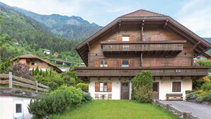 En traditionel træchalet med balkon, omgivet af frodig vegetation og bjerge i baggrunden.