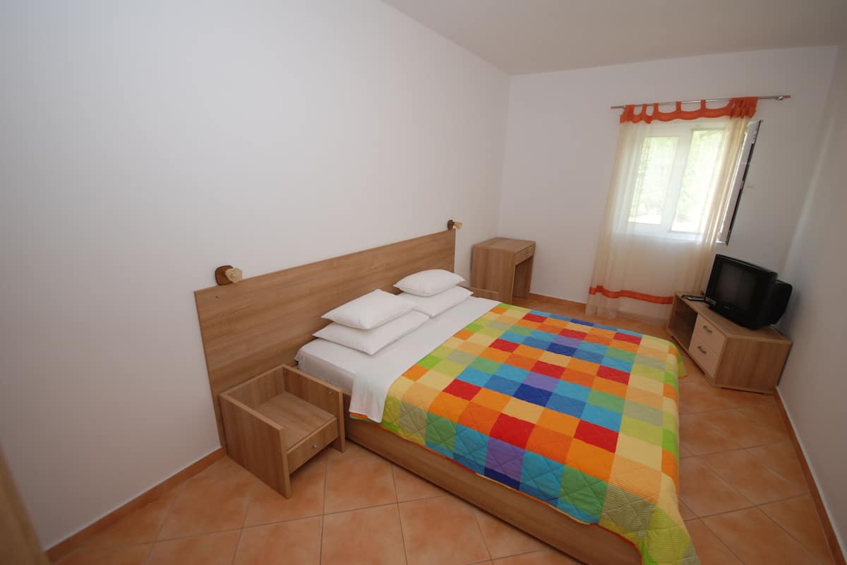 A bedroom with a wooden bed, a colorful quilt, a TV on a wooden stand, and a window with curtains.