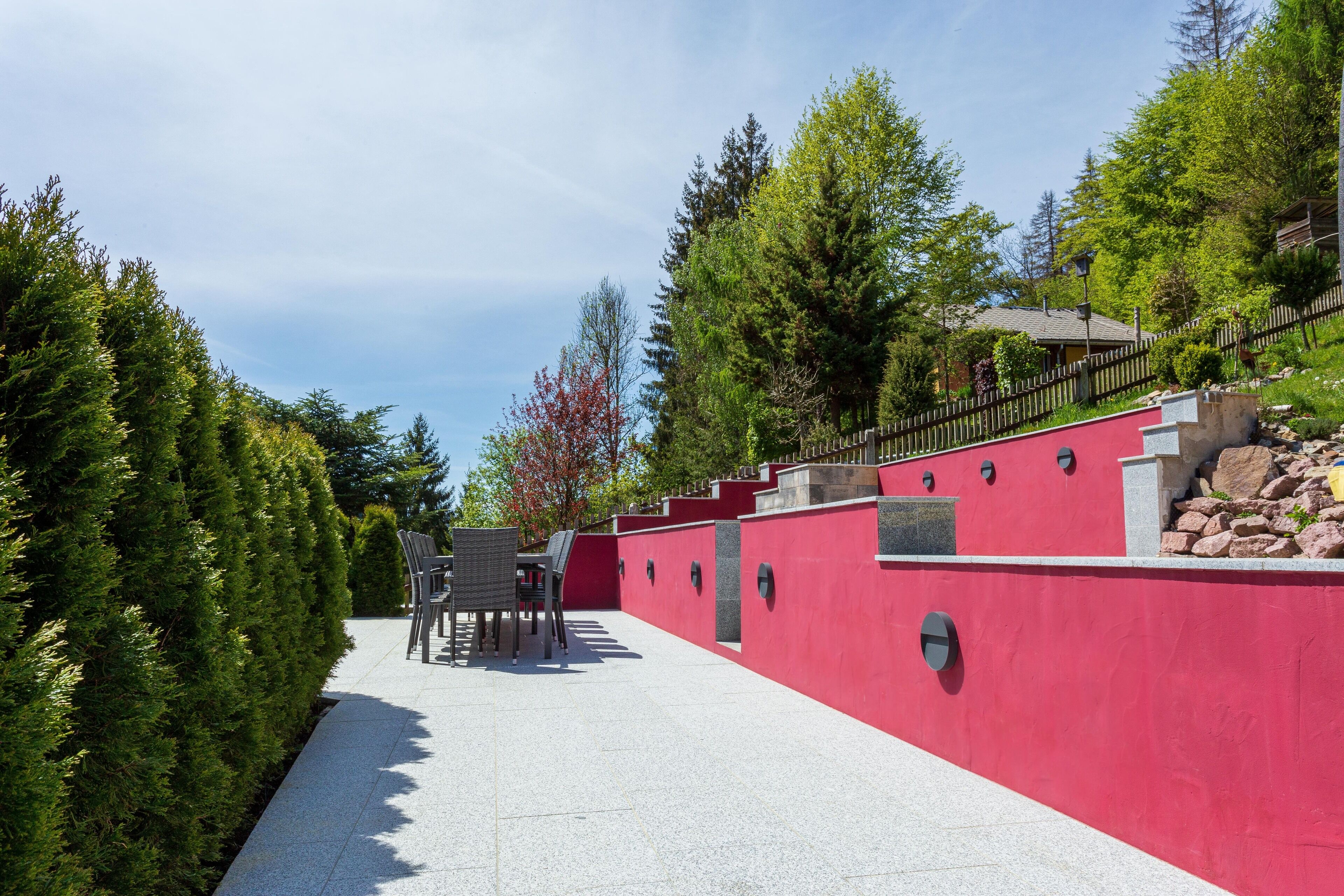 A red wall with a stone bench and a table, surrounded by trees and a clear sky.