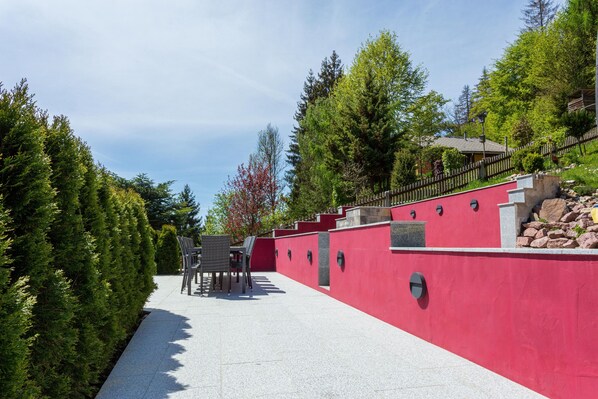 A red wall with a stone bench and a table, surrounded by trees and a clear sky.