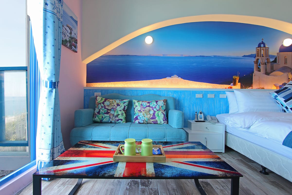 A room with a bed, a sofa, a nightstand, a table with a tray of items, and a mural of a coastal scene.