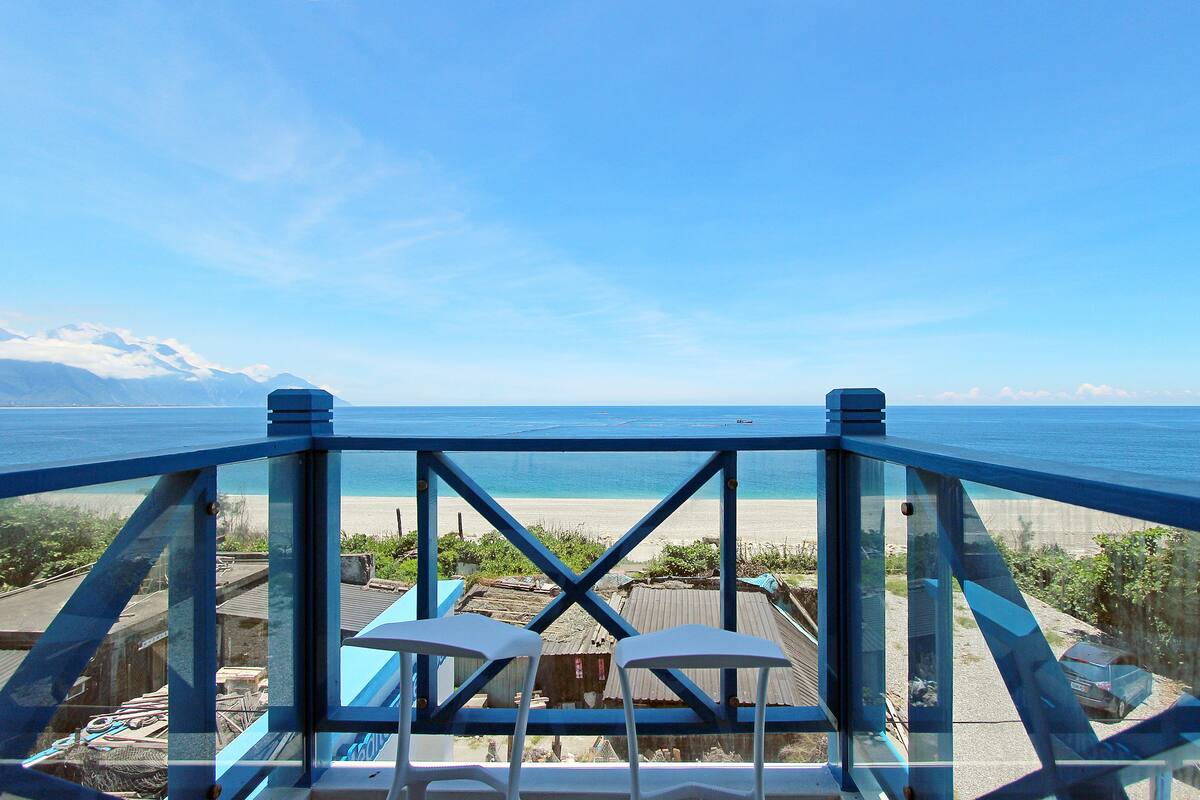 A balcony with two white chairs and a table, overlooking a beach with a clear sky and mountains in the distance.