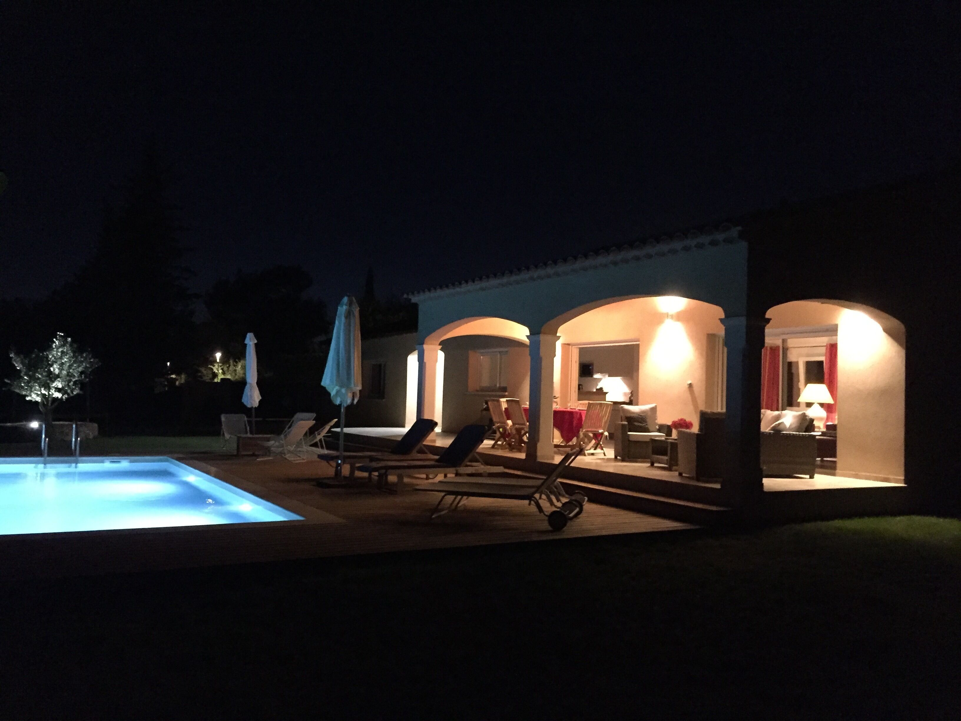 Ein Poolbereich mit Liegestühlen und Sonnenschirmen, ein überdachter Sitzbereich mit Tischen und Stühlen sowie ein gut beleuchteter Innenraum.