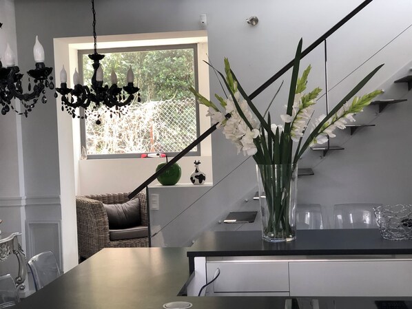 A modern kitchen with a large island, a chandelier, a vase with white flowers, and a view of greenery outside.