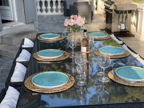 Une table à manger dressée avec des assiettes bleues, des verres transparents et une bouteille de vin, entourée de sets de table en fibres tressées et de coussins blancs.