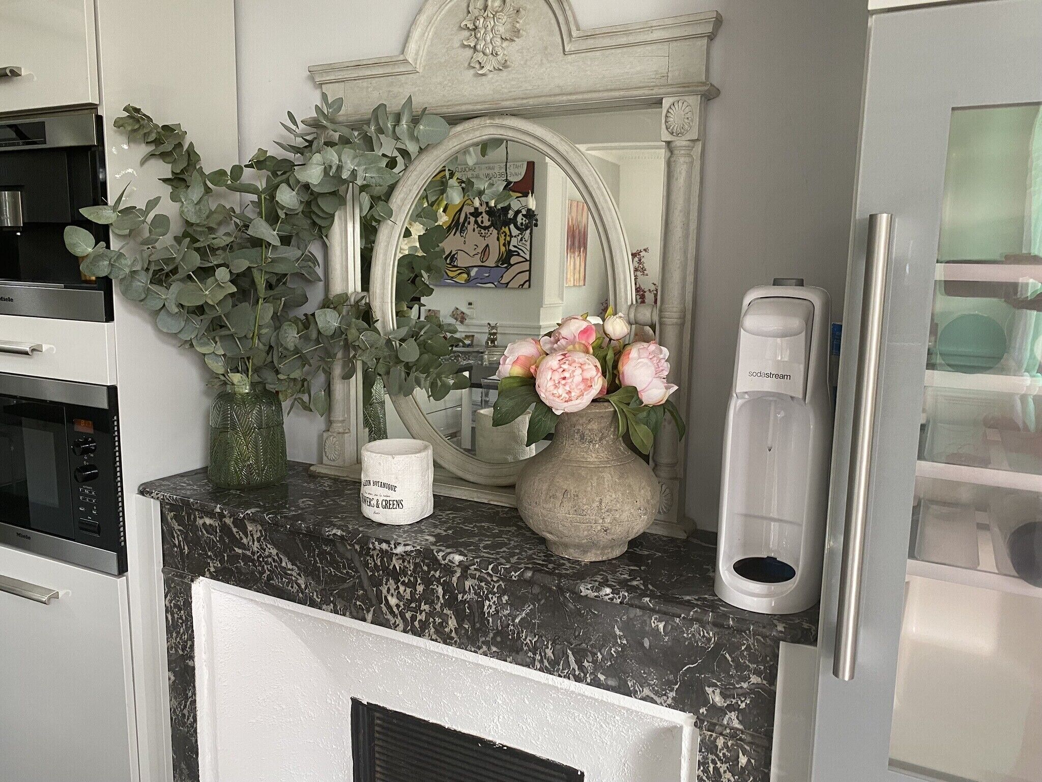 Un comptoir de cuisine surmonté d’un plateau en marbre, un meuble blanc, un grand miroir, un vase avec des fleurs et une machine à café.
